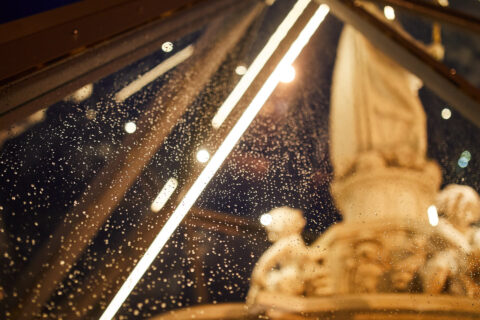 Regentropfen auf der klaren Glasfläche einer beleuchteten Pyramidenstruktur bei Nacht, mit unscharfem barockem Brunnen im Hi…