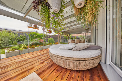 Moderner, barrierefreier Wintergarten mit Glaswänden, Holzboden und Rattan-Lounge-Möbeln, umgeben von einem üppigen Garten i…