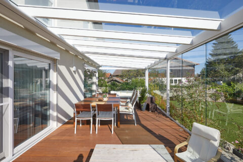 Moderner Kaltwintergarten mit schwellenlosen Glaswänden und Glasdach über einer Holzterrasse, die nahtlos in den Garten über…