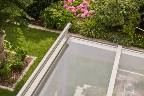 Blick von oben auf einen modernen Wintergarten mit Glasdach und filigraner Konstruktion, umgeben von einem üppigen, grünen G…