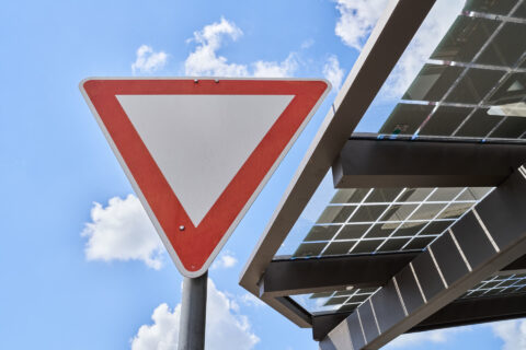 Nahaufnahme eines Vorfahrt-achten-Schilds und einer modernen Terrassenüberdachung mit integrierten Photovoltaikmodulen unter…