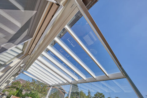 Innenansicht einer Glas-Terrassenüberdachung mit sichtbarer Sparrenkonstruktion und integrierter Markise unter blauem Himmel.
