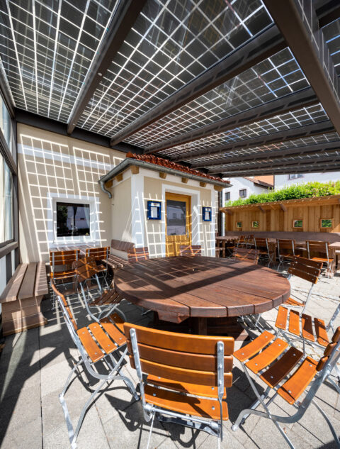Terrassendach mit transparenten Photovoltaik-Modulen über einem Außenbereich mit Holztischen und Stühlen beim Schützenwirt i…