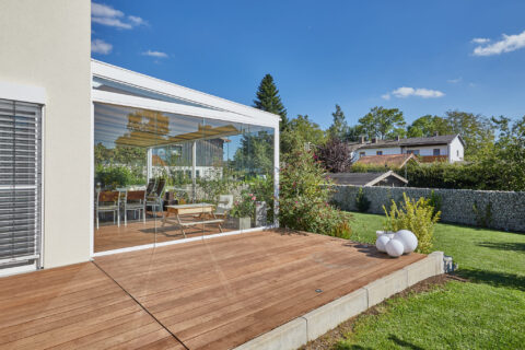 Modernes Kaltwintergarten-System mit rahmenloser Festverglasung, integriert in eine Holzterrasse, mit Blick in den grünen Ga…