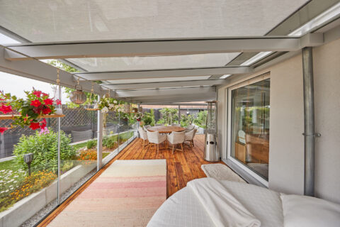 Moderner Kaltwintergarten mit Markise auf einer Holzterrasse in Zorneding, umgeben von Pflanzen und Sitzgelegenheiten.