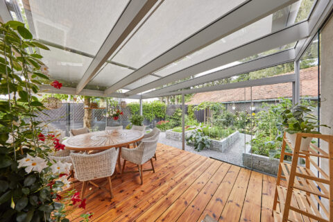 Moderner Ganzglas-Kaltwintergarten mit integrierter Markisenbeschattung, Holzterrasse und bepflanztem Gartenbereich.
