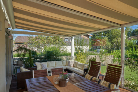 Eine Terrasse mit moderner Ganzglas-Schiebeanlage, gestreifter Markise und bequemen Outdoor-Möbeln, umgeben von einem grünen…
