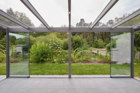 Moderner Kaltwintergarten mit rahmenlosen Glasschiebeelementen und Glasdach, bietet freien Blick in den grünen Garten.