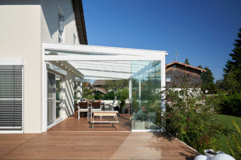 Moderner Kaltwintergarten aus Glas mit seitlichem Windschutz und Glasdach, an einem Einfamilienhaus mit Holzterrasse in sonn…