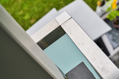 Detailansicht eines Edelstahl-Kantenschutzes an einem Glasgeländer mit Mattglas, Teil eines Balkongeländers.