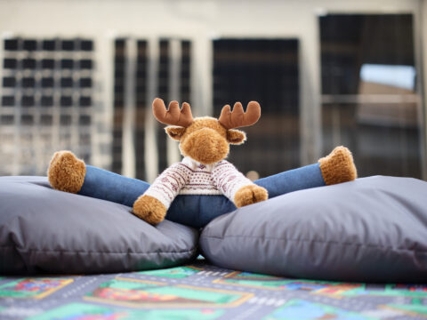 Ein Elch-Stofftier sitzt auf zwei grauen Sitzkissen auf einer bunten Spielmatte, im Hintergrund eine moderne Glasfassade.