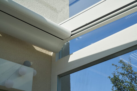 Weißer Markisenkasten integriert in das Gestell eines modernen Kaltwintergartens in Oberpframmern unter blauem Himmel.
