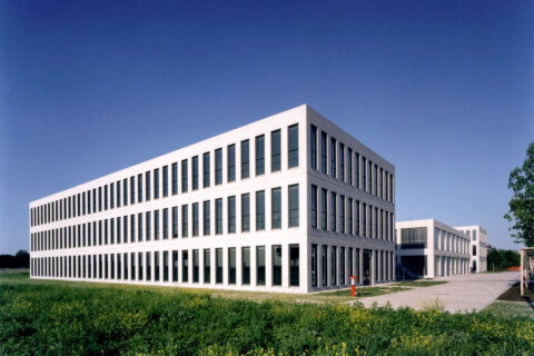 Moderne, dreistöckige Bürogebäude mit heller Metallfassade und vertikalen Fenstern vor blauem Himmel. Umgeben von Grünfläche…