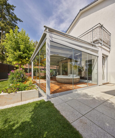 Moderne Glasüberdachung mit Holzterrasse als Wohnraumerweiterung und integrierter Loungemöbel auf sonniger Gartenterrasse in…