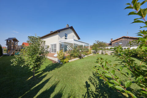 Moderner Kaltwintergarten mit Glasschiebeanlage und Beschattung an einem Einfamilienhaus mit Holzterrasse und Garten in Ober…
