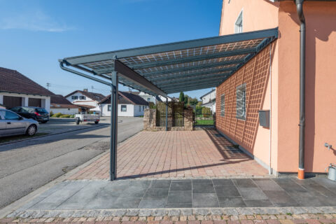 Seitenansicht eines modernen Photovoltaik Carports mit Pultdach und transparenten Glasmodulen vor einem Einfamilienhaus in M…