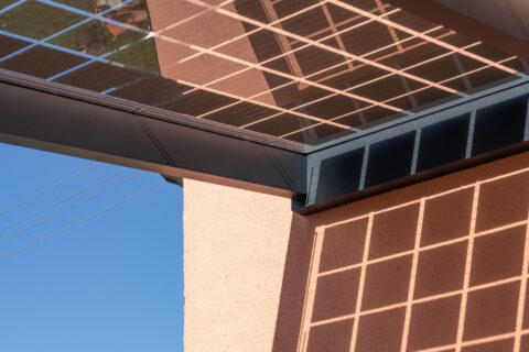 Detailaufnahme eines Photovoltaik-Carports mit Glasmodulen, die rasterförmige Schatten auf eine helle Fassade werfen, blauer…