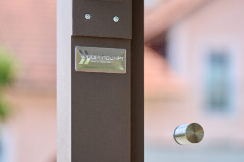 Nahaufnahme eines Glasbaudetails mit Oberhauser Logo auf braunem Profil, Glaselement und Edelstahlhalterung.