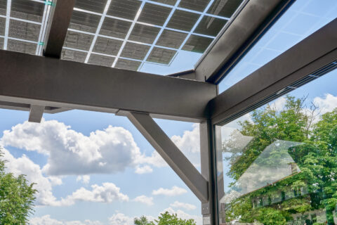 Moderne Terrassenüberdachung aus Glas und dunklem Metall mit integrierten Photovoltaik-Modulen für nachhaltige Energieerzeug…