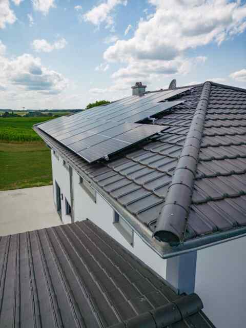 Aufdach-Photovoltaikanlage auf einem modernen Eigenheim mit dunklem Ziegeldach unter blauem Himmel mit weißen Wolken, umgebe…