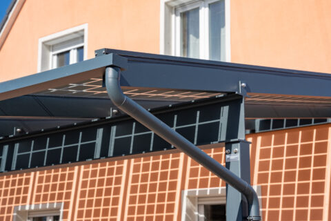 Dunkelgraues PV-Carport aus pulverbeschichtetem Aluminium mit integrierter Regenrinne und Fallrohr, vor einem orangefarbenen…