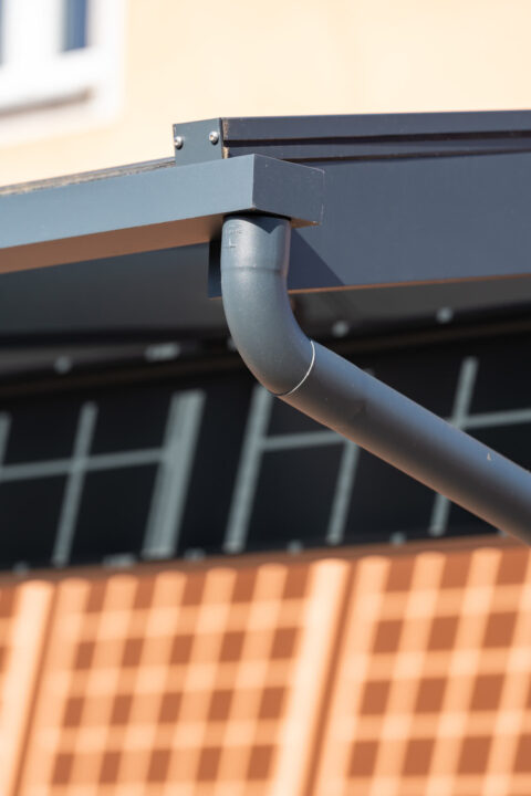 Detailansicht der anthrazitfarbenen Regenrinne eines modernen PV Carports, mit präzisem Schattenwurf auf einer hellbraunen W…