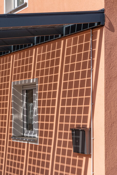 Detailansicht eines Photovoltaik-Moduls, dessen Gitter-Schatten auf eine orangefarbene Hauswand in Mühldorf fällt, mit Fenst…