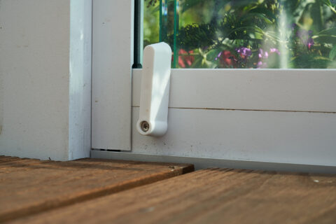 Weißer Griff an Glas-Aluminium-Schiebesystem im Kaltwintergarten. Detailansicht mit Holzterrasse und Blick ins Grüne.