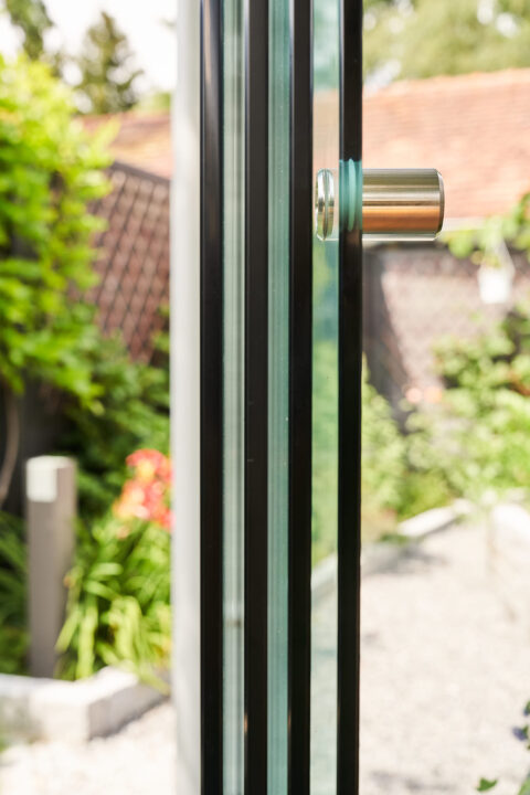 Nahaufnahme eines mehrgleisigen Sunparadise Glasschiebesystems mit schwarzem Profil und Edelstahlgriff, dahinter verschwomme…