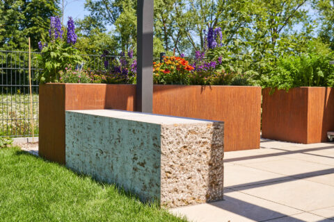 Elegante Terrassendach-Stütze integriert in eine Steinbank, umgeben von Cortenstahl-Pflanzkübeln mit bunten Blumen.