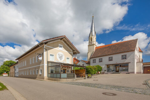 Moderne Glas-Terrassenüberdachung des Gasthofs Schützenwirt in Niederbergkirchen, umgeben von Dorfplatz und Kirche.