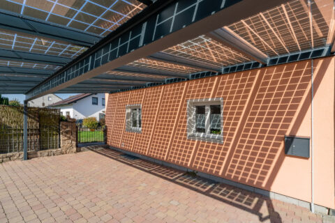Modernes Solarcarport aus pulverbeschichtetem Aluminium mit transparenten Glas-PV-Modulen über Einfahrt eines Einfamilienhau…
