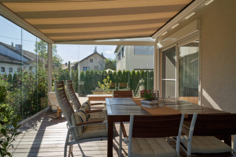 Eleganter Sonnenschutz für eine moderne Terrassenüberdachung aus Glas, mit ausgefahrener Markise und stilvollen Gartenmöbeln.