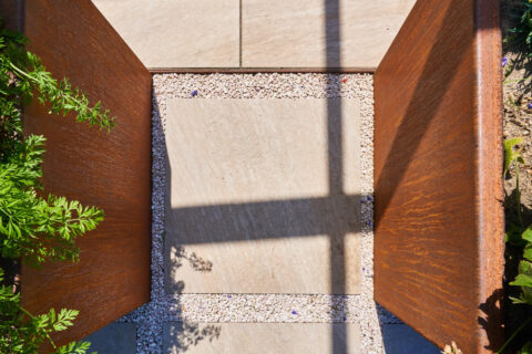 Moderner Terrassenweg aus hellen Platten mit Cortenstahl-Beeteinfassung und Kiesbett, effektvoller Schattenwurf.