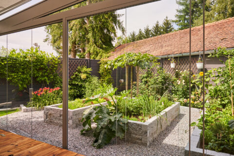 Blick aus einem Wintergarten in Zorneding mit rahmenloser Glas-Schiebeanlage auf einen modernen Terrassengarten mit Hochbeet…