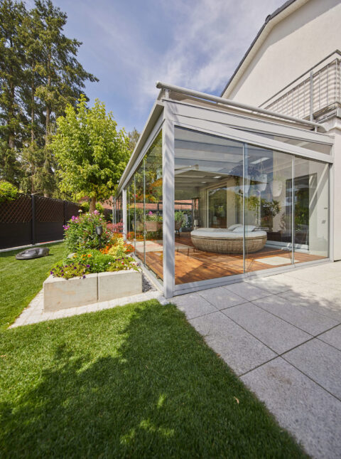 Modernes Glasraum-Terrassendach in Zorneding mit bodentiefen Glaselementen und Holzfußboden, umgeben von einem gepflegten Ga…