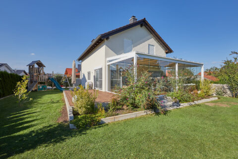 Großzügiger Kaltwintergarten aus Glas am Eigenheim mit Holzterrasse, umgeben von einem grünen Garten und Spielplatz unter bl…