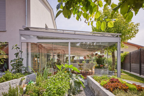 Moderner Glas-Wintergarten an Einfamilienhaus in Zorneding, mit Terrasse, Sitzmöbeln und üppigen Hochbeeten im Vorgarten.
