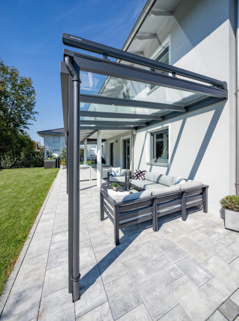 Moderne anthrazitfarbene Aluminium-Terrassenüberdachung mit Glasdach und Aufdachmarkise schützt eine elegante Lounge-Ecke.