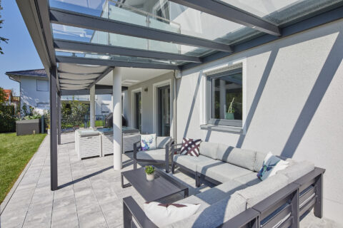Terrassenüberdachung mit Glasdach unter Balkon, moderne Outdoor-Lounge und grauen Fliesen. Ideal für wettergeschützten Aufenthalt.