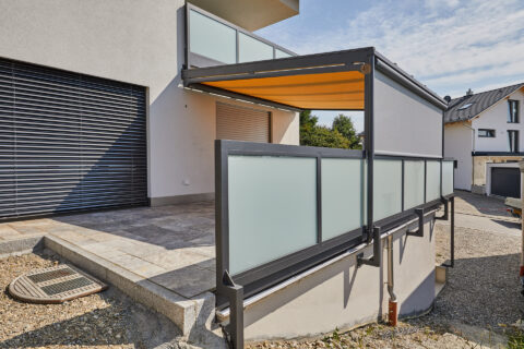 Moderne Terrassenüberdachung mit gelber Unterdachmarkise, grauer Senkrechtmarkise und Glasgeländer an einem Wohnhaus.