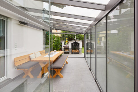 Moderne Glas-Terrassenüberdachung mit Schiebeelementen und Markisen, Blick auf Garten und Außenküche in Kolbermoor.