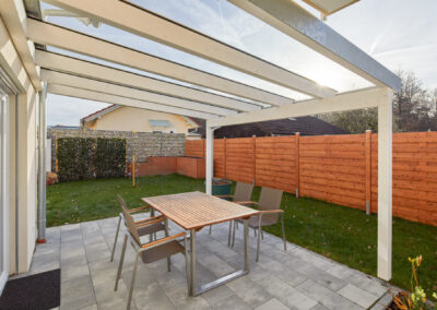 Terrassenüberdachung aus weiß lackiertem Holz mit transparentem Glasdach über einer modernen Steinterrasse mit Gartenmöbeln.