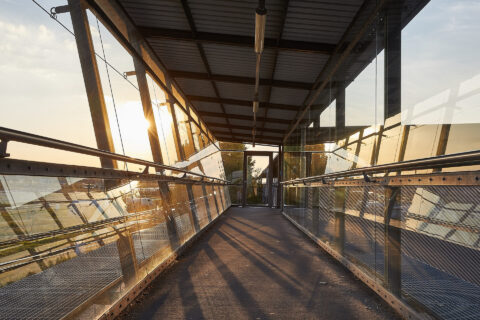 Moderne transparente Glasbrücke mit Stahlkonstruktion am Bahnhof Mühldorf am Inn bei Sonnenuntergang, blendendes Licht und S…