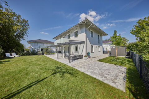 Moderne Terrassenüberdachung in Dunkelgrau an einem hellgrauen Einfamilienhaus mit stilvollen Loungemöbeln und gepflegtem Garten.