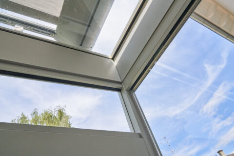 Nahaufnahme einer modernen Glas-Wintergarten-Dachkonstruktion mit integrierter Beschattungsanlage, Blick auf Himmel und Bäum…