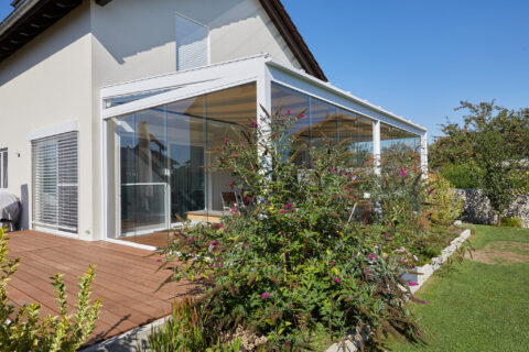 Wetterfester Kaltwintergarten aus Glas und Aluminium an einem Einfamilienhaus mit Holzterrasse und Garten.