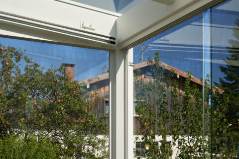 Detailansicht eines Kaltwintergartens mit weißem Rahmen und klarem Glas, das einen Garten mit Apfelbaum und Holzhaus zeigt.