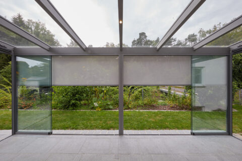 Moderner Kaltwintergarten aus Glas mit integriertem, ausfahrbarem Sonnenschutz und Blick auf den grünen Garten.