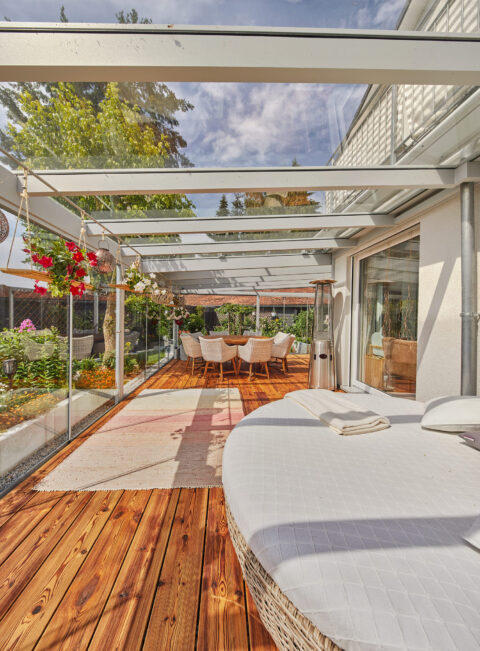 Moderne Ganzglas-Terrassenüberdachung mit Holzboden, Loungemöbeln und Essbereich, umgeben von Garten und Pflanzen in Zorneding